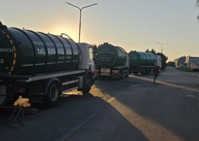 Trzy zielone ciężarówki-cysterny stojące w rzędzie na drodze, prawdopodobnie o zachodzie słońca, tworząc długie cienie. W tle widać latarnie uliczne i niewyraźne drzewa.