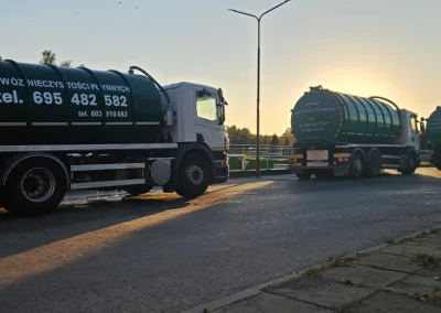 Trzy ciężarówki-cysterny w kolorze ciemnozielonym stojące w rzędzie na drodze, z widocznym wschodzącym lub zachodzącym słońcem w tle, które tworzy długie cienie. Na boku najbliższej cysterny widnieje napis "WYWÓZ NIECZYSTOŚCI PŁYNNYCH" oraz numery telefonów: "tel. 695 482 582" i "tel. 603 610 682".