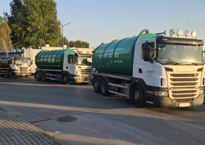 Trzy białe ciężarówki Scania z zielonymi cysternami stoją w rzędzie na asfaltowej drodze, prawdopodobnie czekając na rozładunek lub załadunek.