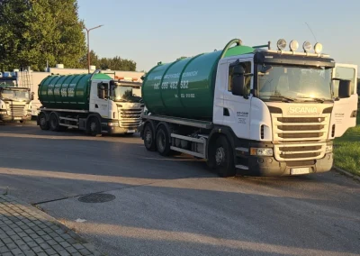 Trzy białe ciężarówki Scania z zielonymi cysternami stoją w rzędzie na asfaltowej drodze, prawdopodobnie czekając na rozładunek lub załadunek. Widoczne są numery telefonów na boku jednej z cystern.